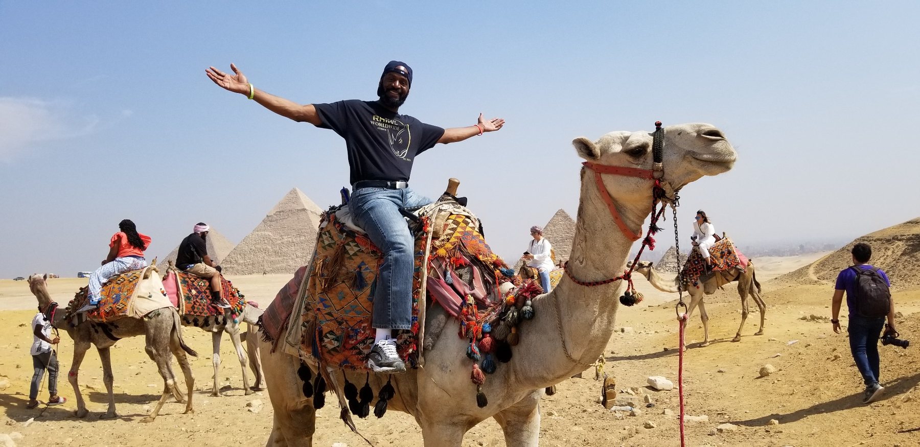 Darnell riding a camel with arms open in front of the pyramids